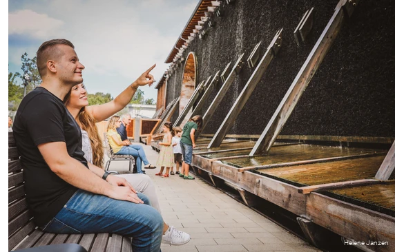 Ein junges Pärchen sitzt auf einer Bank am Gradierwerk im Kurpark Bad Sassendorf. Sie zeigt mit dem Finger in Richtung Gradierwerk und erläutert, warum die Sole am Gradierwerk herunterläuft.