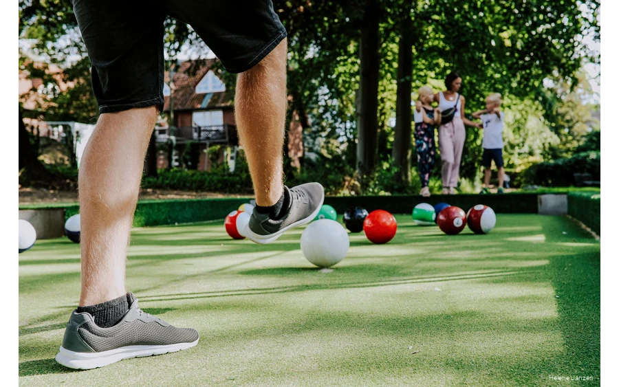 Auf dem Fußballbillardfeld liegen verschieden farbige Fußbälle, die wie große Billardkugeln aussehen. Ein junger Mann schießt dem weißen Ball in Richtung des roten Balls. Seine Frau und seine Kinder (ein Junge und ein Mädchen) schauen ihm beim Spiel zu.
