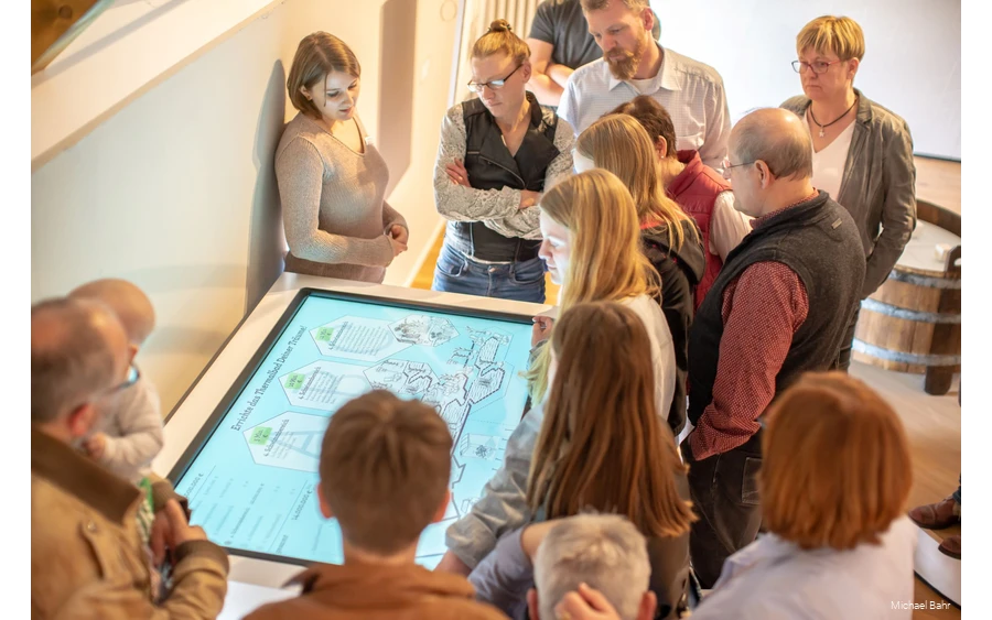 Eine Gruppe steht im Museum vor einem Bildschirm, ein Museumsguide erklärt etwas.