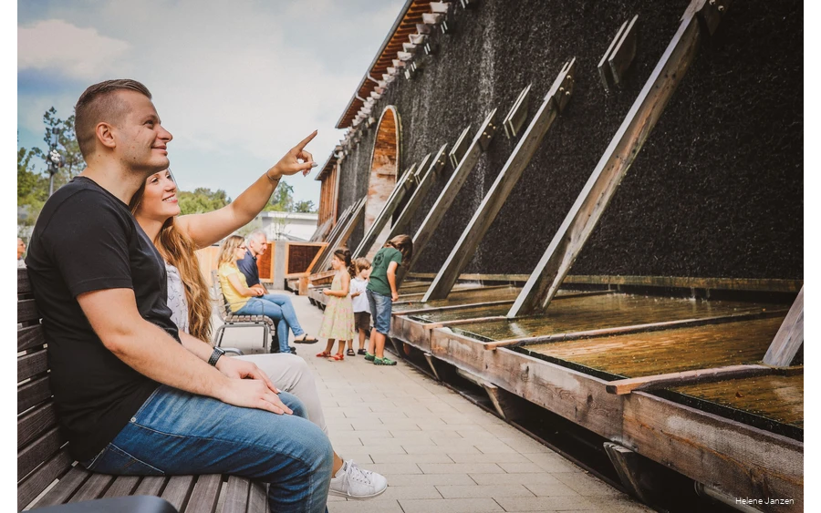 Ein Pärchen sitzt auf einer Bank vor dem Gradierwerk. Sie zeigt auf das Gradierwerk und erzählt ihrem Freund etwas.