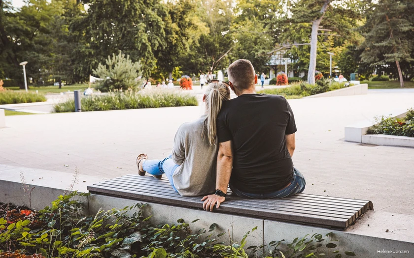 Ein junges Pärchen sitzt auf einer Bank im Kurpark. Drumherum sind schöne Blumenbeete angelegt. Leute spazieren im Kurpark.