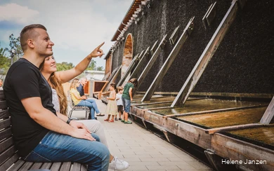 Ein junges Pärchen sitzt auf einer Bank am Gradierwerk im Kurpark Bad Sassendorf. Sie zeigt mit dem Finger in Richtung Gradierwerk und erläutert, warum die Sole am Gradierwerk herunterläuft.