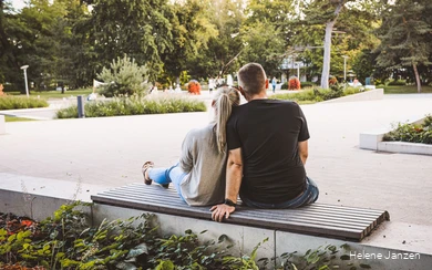 Ein junges Pärchen sitzt auf einer Bank im Kurpark. Drumherum sind schöne Blumenbeete angelegt. Leute spazieren im Kurpark.