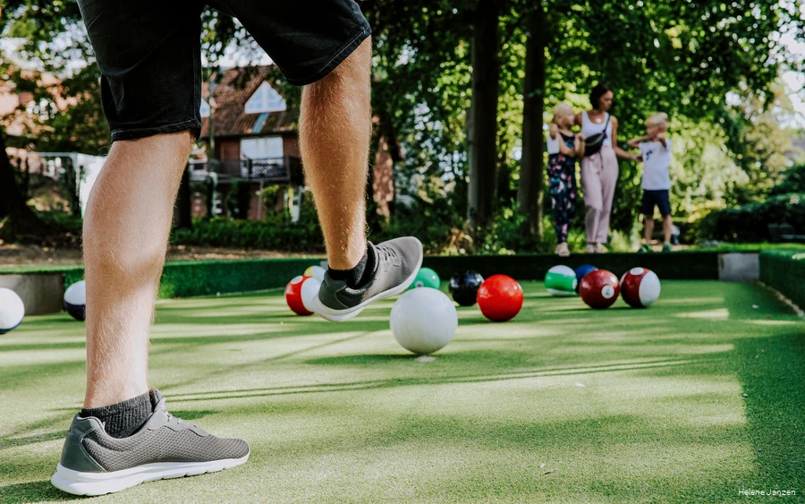 Auf dem Fußballbillardfeld liegen verschieden farbige Fußbälle, die wie große Billardkugeln aussehen. Ein junger Mann schießt dem weißen Ball in Richtung des roten Balls. Seine Frau und seine Kinder (ein Junge und ein Mädchen) schauen ihm beim Spiel zu.