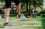 Auf dem Fußballbillardfeld liegen verschieden farbige Fußbälle, die wie große Billardkugeln aussehen. Ein junger Mann schießt dem weißen Ball in Richtung des roten Balls. Seine Frau und seine Kinder (ein Junge und ein Mädchen) schauen ihm beim Spiel zu.
