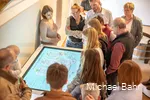 Eine Gruppe steht im Museum vor einem Bildschirm, ein Museumsguide erklärt etwas.